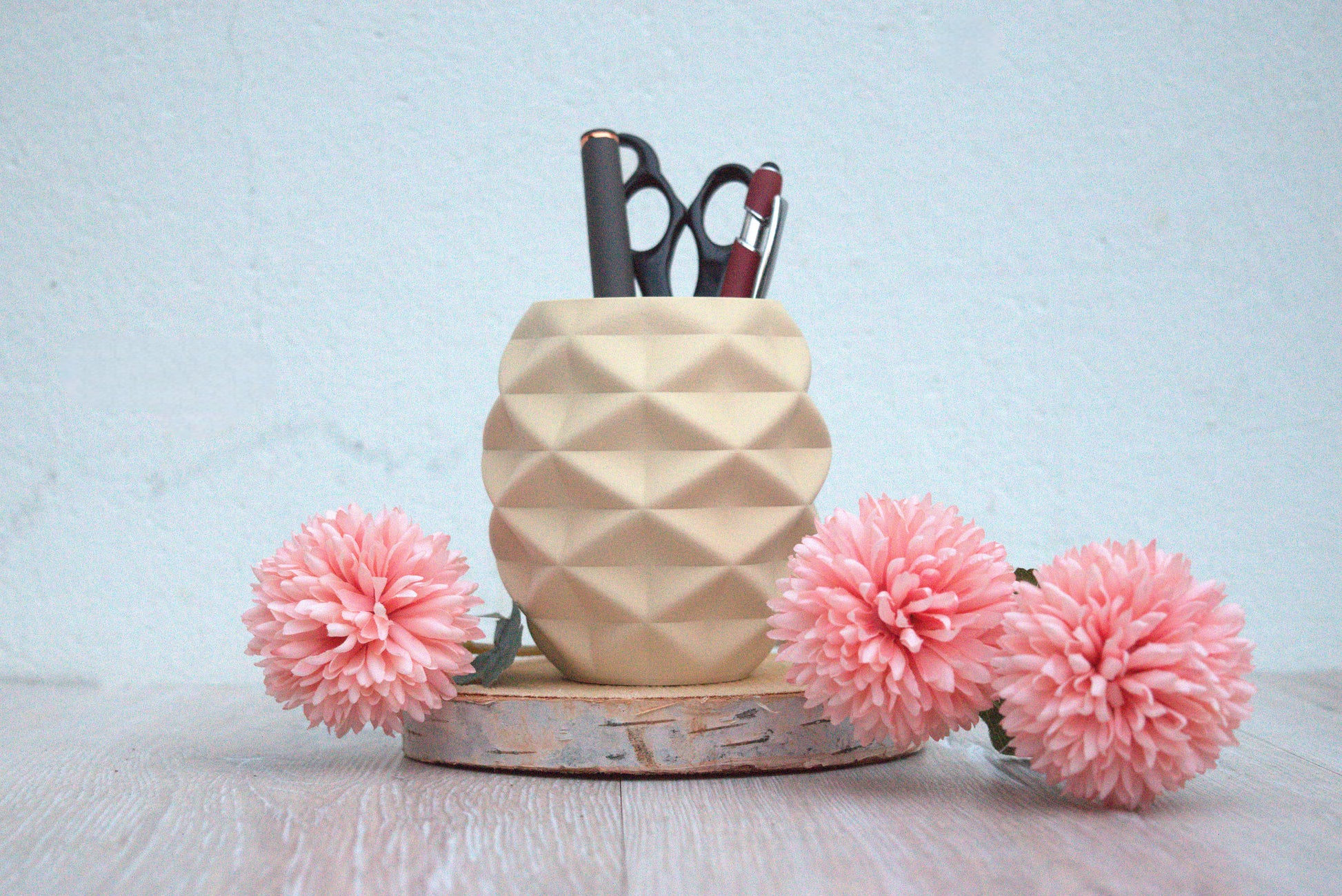 Drei pinke, runde Kunstblumen umrahmen einen geometrischen Behältnis (Stiftehalter) in beige (Weißeiche), der auf einem Stück Rinde platziert ist. Innerhalb des Behälters liegen ein schlanker Stift und eine kleine Schere.
Besteht aus PLA Wood.