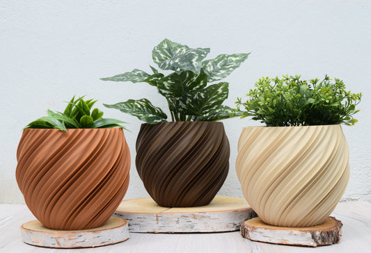 Drei moderne Pflanzgefäße mit gedrehtem Spiraldesign in Terrakotta (Lehmbraun), Dunkelbraun (Schwarze Walnuss) und beige (Weißeiche) präsentieren verschiedene grüne Pflanzen auf rustikalen, runden Holzuntersetzern.

Sie haben einen Innentopf mit Ablauflöchern. der Innentopf ist schwarz.